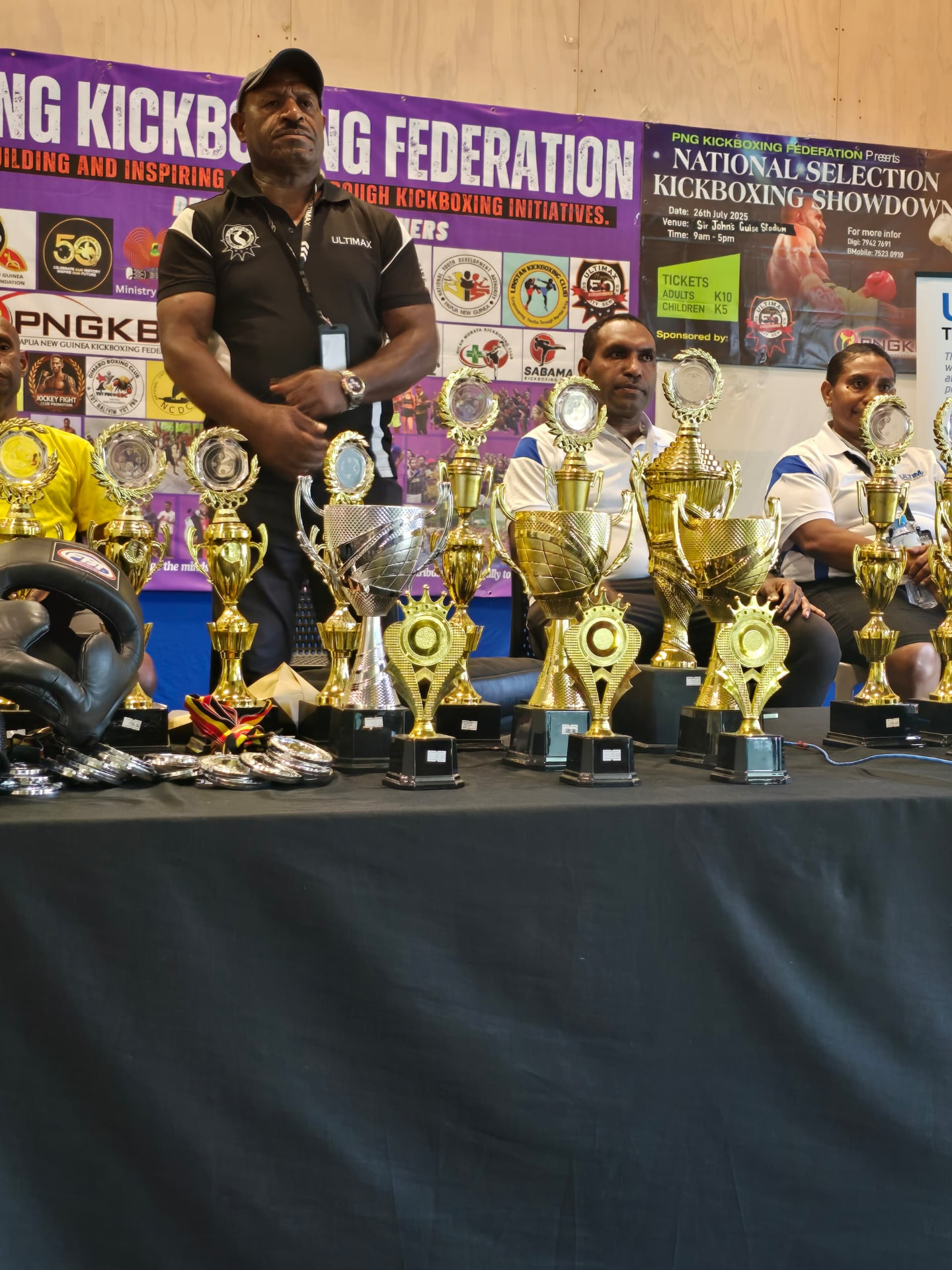 PNGKBF officials seated behind rows of gold trophies under the federation banner