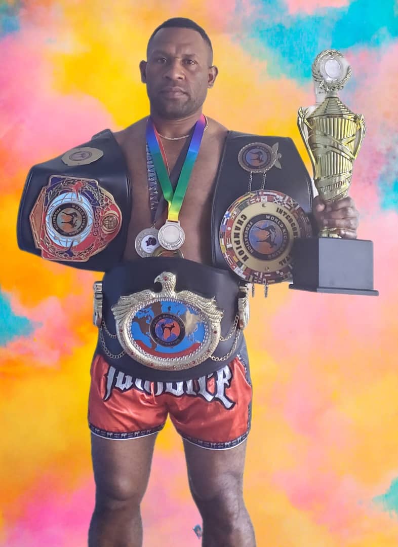 PNG kickboxing world champion holding three championship belts, medals and a gold trophy